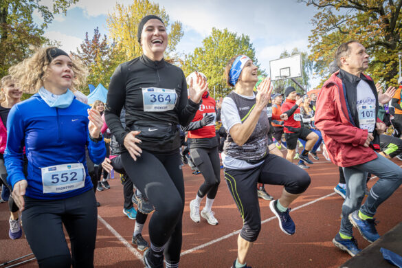Gromada uśmiechniętych biegaczy na czerwonej bieżni podczas rozgrzewki. Ludzie w kolorowych strojach sportowych z numerami startowymi radośnie podskakują.