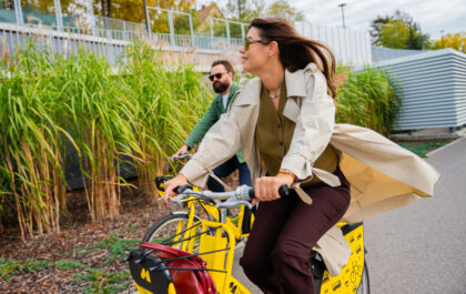 Para jedzie na żółtych rowerach miejskich Metrorower wzdłuż zielonych krzewów. Kobieta w beżowym płaszczu uśmiecha się, a mężczyzna jedzie za nią.