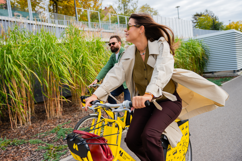 Para jedzie na żółtych rowerach miejskich Metrorower wzdłuż zielonych krzewów. Kobieta w beżowym płaszczu uśmiecha się, a mężczyzna jedzie za nią.