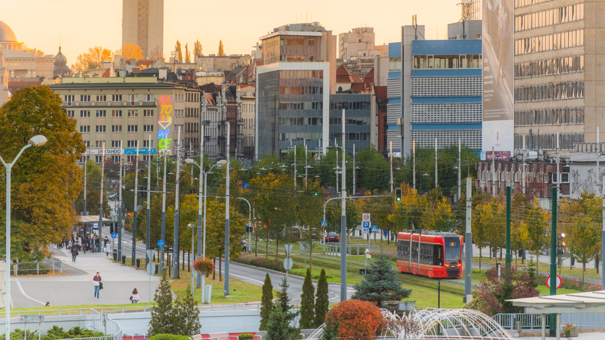 Widok na centrum Katowic o zachodzie słońca. Czerwony tramwaj mknie obok fontanny, w tle widać bloki i kolorowy neon z nazwą miasta. Jest jesiennie.