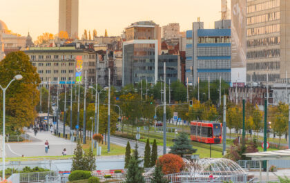 Widok na centrum Katowic o zachodzie słońca. Czerwony tramwaj mknie obok fontanny, w tle widać bloki i kolorowy neon z nazwą miasta. Jest jesiennie.