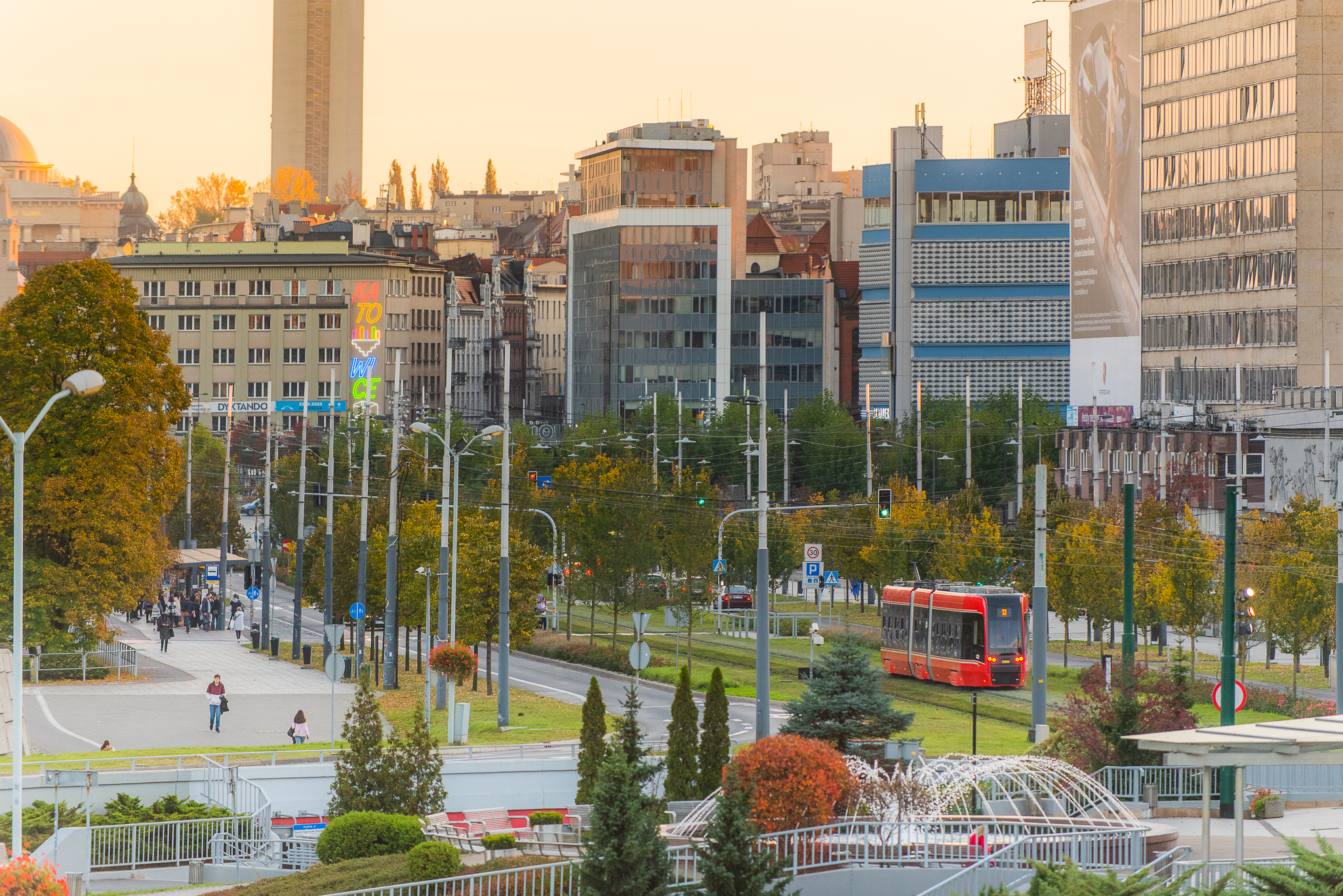 Widok na centrum Katowic o zachodzie słońca. Czerwony tramwaj mknie obok fontanny, w tle widać bloki i kolorowy neon z nazwą miasta. Jest jesiennie.