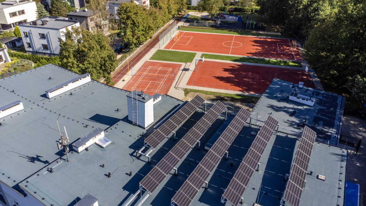 Widok z góry na płaski dach budynku z panelami fotowoltaicznymi. W tle wielofunkcyjne boiska sportowe oraz osiedle domów jednorodzinnych w Katowicach.