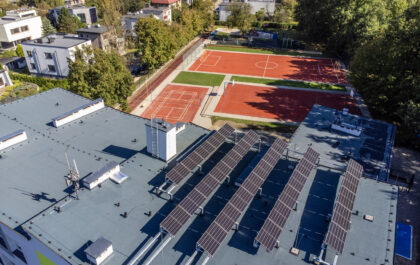 Widok z góry na płaski dach budynku z panelami fotowoltaicznymi. W tle wielofunkcyjne boiska sportowe oraz osiedle domów jednorodzinnych w Katowicach.