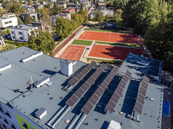 Widok z góry na płaski dach budynku z panelami fotowoltaicznymi. W tle wielofunkcyjne boiska sportowe oraz osiedle domów jednorodzinnych w Katowicach.