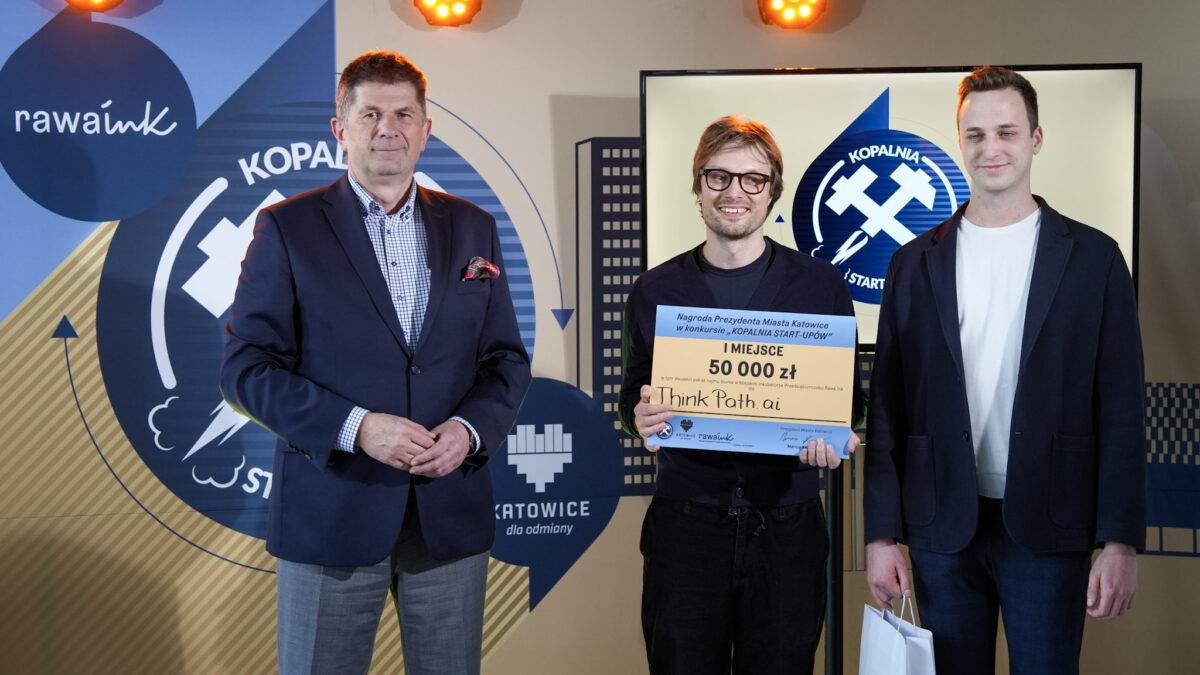 Trzej mężczyźni stoją na tle logotypów Rawa.Ink i Katowic. Środkowy trzyma symboliczny czek na 50 000 zł za I miejsce w konkursie Kopalnia Start-upów.