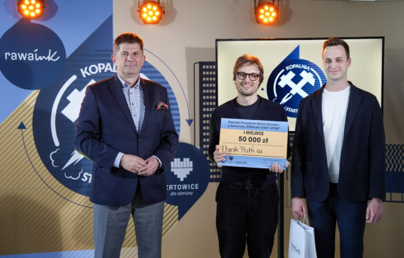 Trzej mężczyźni stoją na tle logotypów Rawa.Ink i Katowic. Środkowy trzyma symboliczny czek na 50 000 zł za I miejsce w konkursie Kopalnia Start-upów.