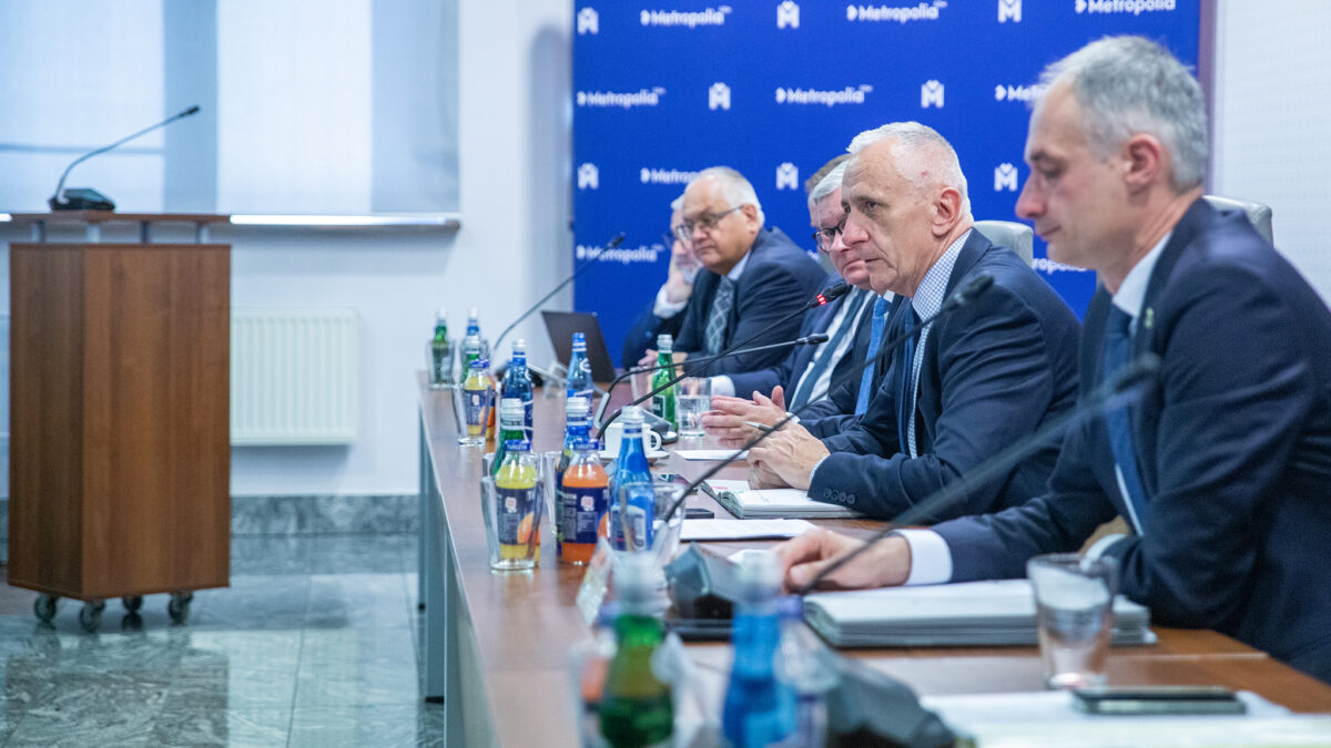 Zdjęcie przedstawia pięciu mężczyzn w garniturach siedzących przy długim stole prezydialnym na tle niebieskiej ścianki z białym logo „Metropolia”.