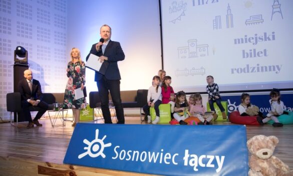 Scena z napisem „Sosnowiec łączy”. Mężczyzna mówi do mikrofonu, obok stoją dorośli, a z prawej na pufach siedzą dzieci. W tle ekran.