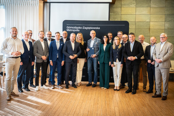 Na zdjęciu widoczna jest grupa osób w strojach biznesowych, stojących przed czarnym tłem z logo Górnośląsko-Zagłębiowskiej Metropolii. Część osób pozuje, trzymając dokument w teczce.