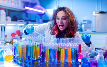 Zbliżenie na podekscytowaną, młodą kobietę w laboratorium, pracującą przy stanowisku pełnym probówek z kolorowymi płynami. Ma na sobie biały fartuch, okulary ochronne i niebieskie rękawiczki. Trzyma pipetę w jednej ręce i zagląda w stronę probówek z otwartymi z wrażenia ustami. Włosy ma kręcone, z kolorowymi pasmami (różowe i pomarańczowe). Tło to typowe wyposażenie laboratorium w chłodnym świetle.